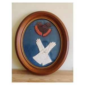 May include: A framed oval picture with a blue background. The picture features a pair of white gloves and a pair of brown leather gloves with orange trim.