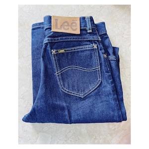 May include: Dark blue denim jeans with a classic five-pocket design. The jeans feature a leather "Lee" brand patch and yellow stitching. The jeans are folded neatly, showcasing the back pocket and the brand label.