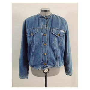 May include: A blue denim jacket with a mandarin collar and button closure. The jacket has two front pockets and a single pocket on the left chest.