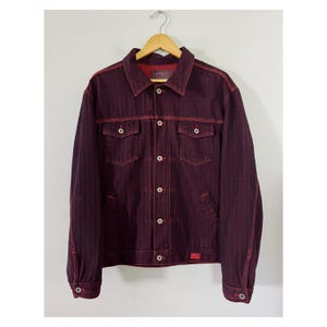 May include: A deep burgundy denim jacket featuring a classic collar, long sleeves with buttoned cuffs, and a front button closure. The garment includes two chest pockets with button flaps, two side pockets, and contrasting red stitching throughout.