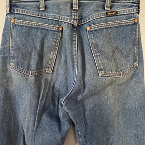 Puede incluir: Vaqueros de mezclilla azul con dise&ntilde;o de cintura alta, trabillas para cintur&oacute;n, dos bolsillos traseros con remaches de color cobre y una etiqueta negra de la marca Wrangler cosida sobre el bolsillo derecho. El tejido tiene un lavado medio.