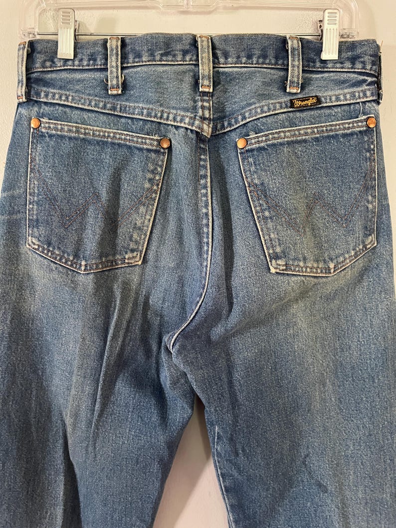 Puede incluir: Vaqueros de mezclilla azul con dise&ntilde;o de cintura alta, trabillas para cintur&oacute;n, dos bolsillos traseros con remaches de color cobre y una etiqueta negra de la marca Wrangler cosida sobre el bolsillo derecho. El tejido tiene un lavado medio.