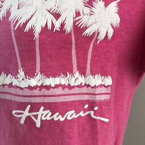 May include: Pink t-shirt with a white graphic of palm trees and the word "Hawaii" printed on the front.