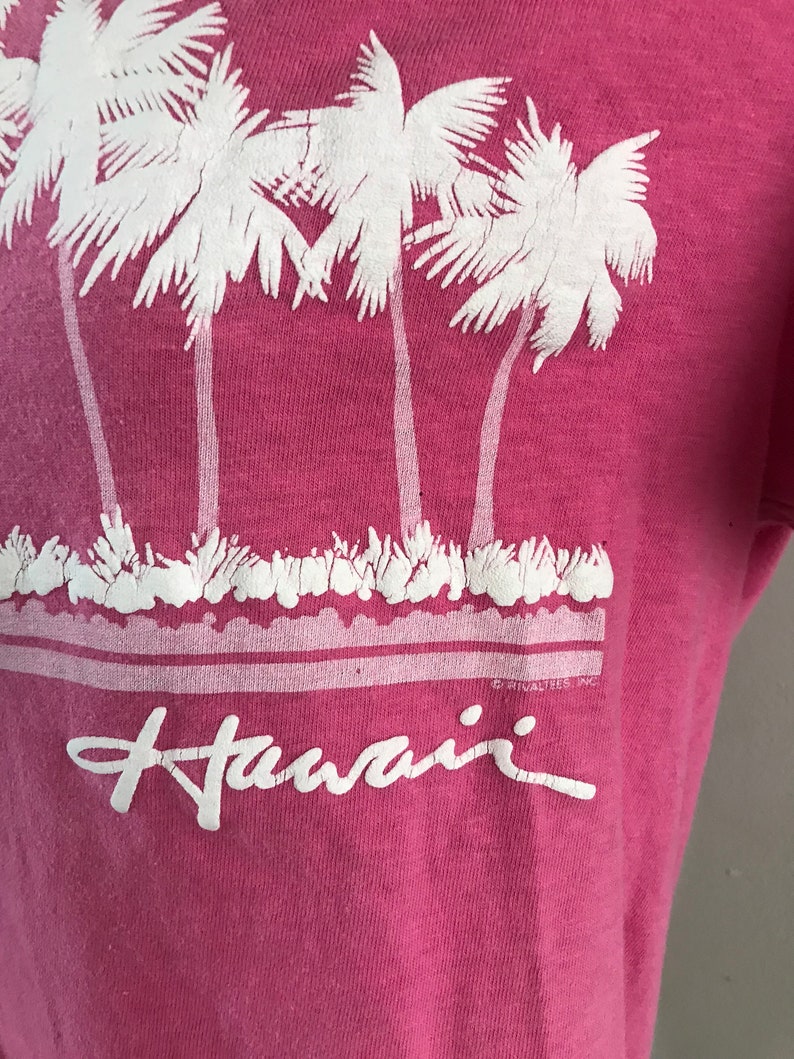 May include: Pink t-shirt with a white graphic of palm trees and the word "Hawaii" printed on the front.