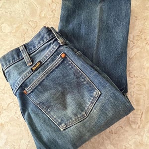 Puede incluir: Un par de vaqueros de mezclilla en tono azul medio con un dise&ntilde;o cl&aacute;sico de cinco bolsillos, cierre de botones y trabillas para cintur&oacute;n. El bolsillo trasero muestra una etiqueta negra de la marca Wrangler con letras doradas y remaches.