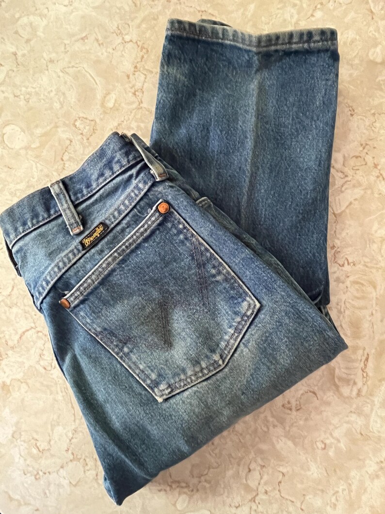 Puede incluir: Un par de vaqueros de mezclilla en tono azul medio con un dise&ntilde;o cl&aacute;sico de cinco bolsillos, cierre de botones y trabillas para cintur&oacute;n. El bolsillo trasero muestra una etiqueta negra de la marca Wrangler con letras doradas y remaches.