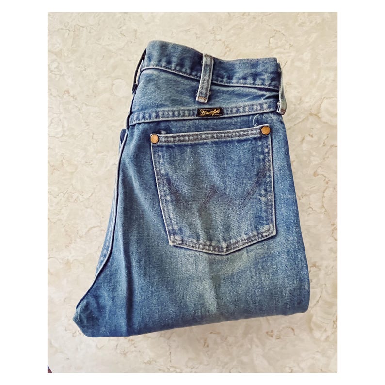 Puede incluir: Unos vaqueros de mezclilla azul con un dise&ntilde;o cl&aacute;sico de cinco bolsillos, cintura alta con trabillas para cintur&oacute;n y una etiqueta de la marca Wrangler sobre el bolsillo trasero. Los vaqueros tienen remaches de color cobre y una W cosida.