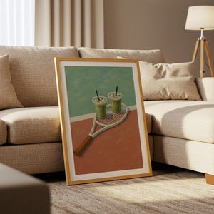 以下が含まれることがあります： テニスラケットと2つの緑色の飲み物が描かれた額入りのアートプリント。背景はターコイズと赤の配色です。アートワークは、リビングルームのベージュのソファに展示されています。フレームは木製です。