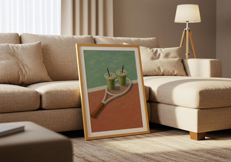 以下が含まれることがあります： テニスラケットと2つの緑色の飲み物が描かれた額入りのアートプリント。背景はターコイズと赤の配色です。アートワークは、リビングルームのベージュのソファに展示されています。フレームは木製です。