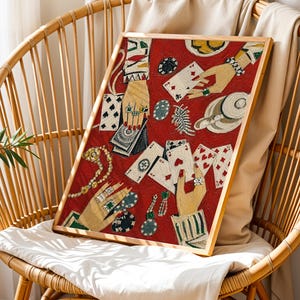 K&ouml;nnte beinhalten: Ein gerahmtes Kunstwerk mit rotem Hintergrund, das Spielkarten, mit Schmuck geschm&uuml;ckte H&auml;nde und Pokerchips zeigt. Das Kunstwerk wird auf einem Korbstuhl mit wei&szlig;em Kissen und beigem &Uuml;berwurf pr&auml;sentiert.