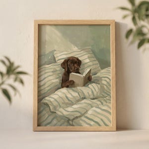 May include: A framed print featuring a brown puppy reading a book while nestled in bed. The bedding and pillows have a striped pattern in shades of blue and white. The frame is made of light-colored wood.