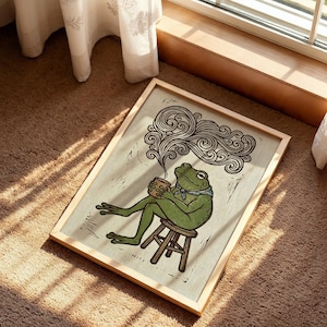 May include: A framed art print depicting a green frog seated on a stool, holding a cup, and enjoying a hot drink. The frog wears a blue scarf. The artwork is in a woodcut style, with swirling steam rising from the cup.