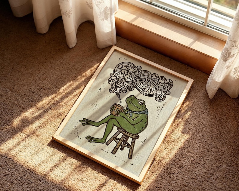 May include: A framed art print depicting a green frog seated on a stool, holding a cup, and enjoying a hot drink. The frog wears a blue scarf. The artwork is in a woodcut style, with swirling steam rising from the cup.