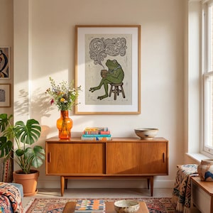 May include: A framed art print shows a green frog on a stool, holding a mug with steam. The artwork is in a wooden frame, above a wooden cabinet with an orange vase and plants. The room has a warm atmosphere, with a rug and a window.