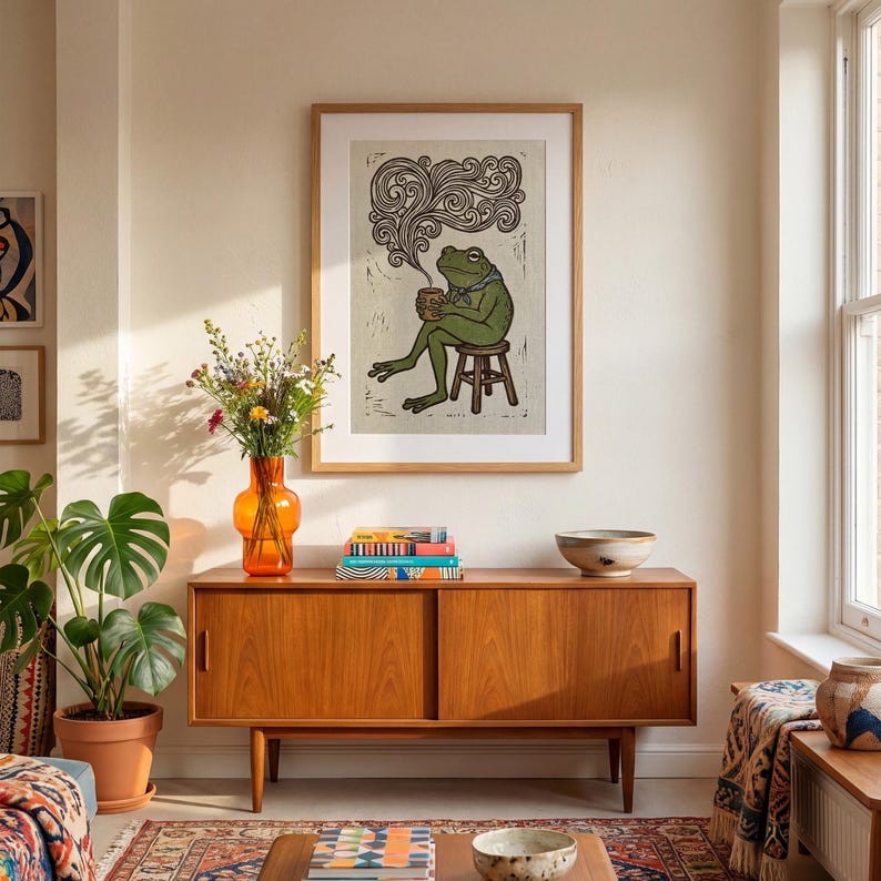 May include: A framed art print shows a green frog on a stool, holding a mug with steam. The artwork is in a wooden frame, above a wooden cabinet with an orange vase and plants. The room has a warm atmosphere, with a rug and a window.