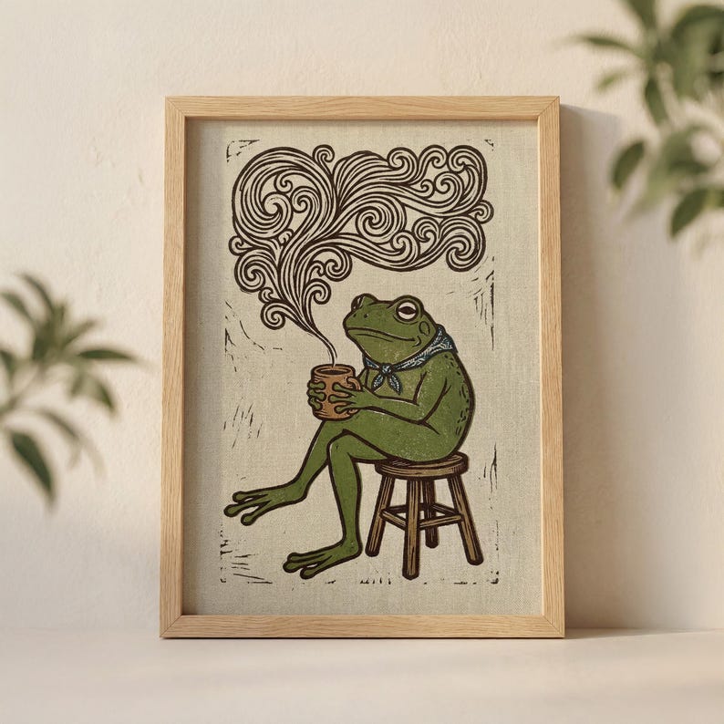 May include: A framed art print depicting a green frog seated on a stool, holding a cup, and exhaling steam. The frog wears a blue bandana. The print is in a woodcut style with brown and beige tones.