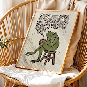 May include: A framed art print depicting a green frog seated on a stool, holding a mug, with steam rising. The frog wears a blue neckerchief. The artwork has a rustic, linocut style with a light beige background and a wooden frame.