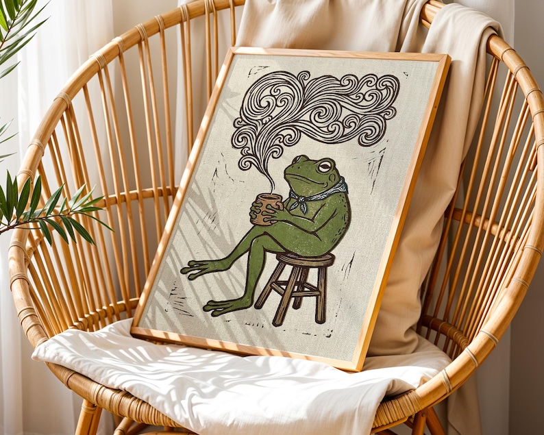 May include: A framed art print depicting a green frog seated on a stool, holding a mug, with steam rising. The frog wears a blue neckerchief. The artwork has a rustic, linocut style with a light beige background and a wooden frame.