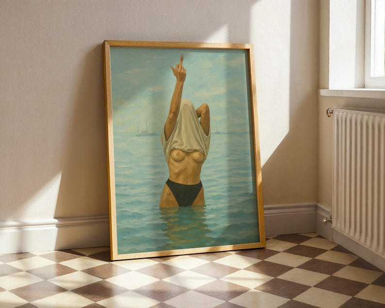 Puede incluir: Impresi&oacute;n art&iacute;stica enmarcada que representa una figura en el agua, parcialmente oculta por una camiseta blanca que se sube por la cabeza. La figura lleva una parte inferior negra, con el dedo coraz&oacute;n levantado. El fondo representa un paisaje marino con veleros.