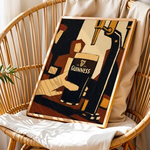 Op de afbeelding: Een ingelijste kunstafdruk met een Guinness-advertentie. Het kunstwerk toont een hand die een pint Guinness inschenkt, met het logo van het merk. Het kleurenpalet omvat zwart-, beige- en bruintinten, met een houten lijst.