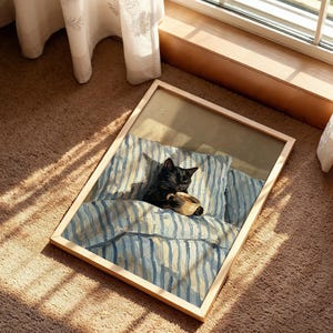 May include: A framed artwork depicts a black cat nestled in a bed with blue and white striped bedding, holding a cup. The artwork is in a light wooden frame, resting on a brown carpet near a window with sheer curtains.