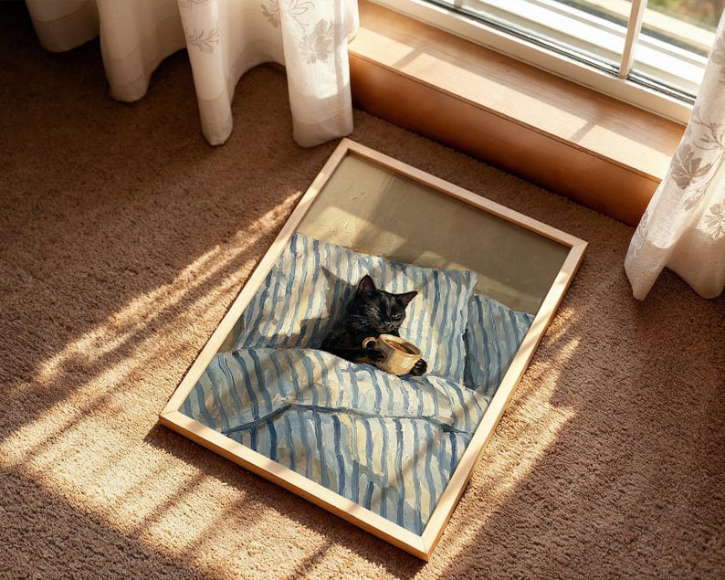 May include: A framed artwork depicts a black cat nestled in a bed with blue and white striped bedding, holding a cup. The artwork is in a light wooden frame, resting on a brown carpet near a window with sheer curtains.