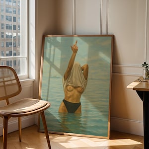 Puede incluir: Una obra de arte enmarcada representa una figura en el agua, levantando una mano con el dedo medio extendido. La figura est&aacute; parcialmente oculta por una camiseta blanca. La obra est&aacute; en un marco de madera, apoyada contra una pared cerca de una ventana y una silla de madera.