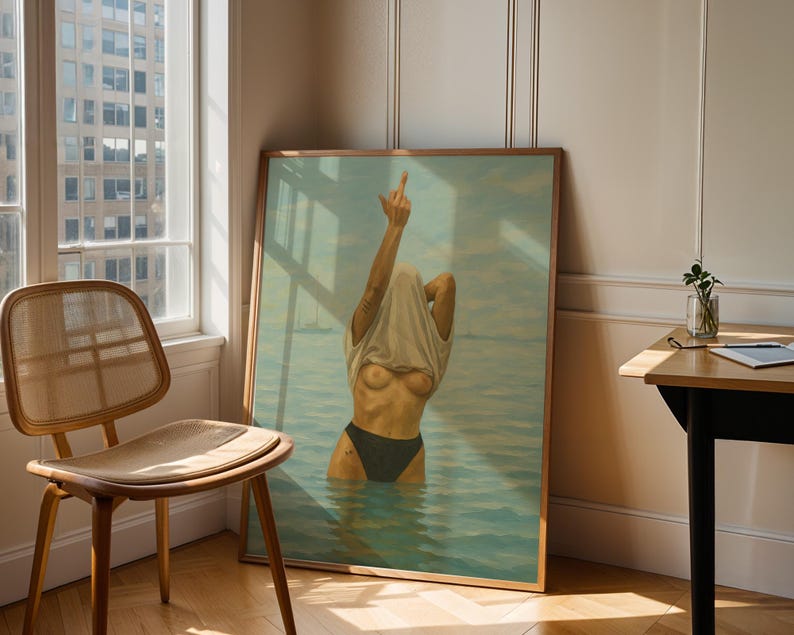 Puede incluir: Una obra de arte enmarcada representa una figura en el agua, levantando una mano con el dedo medio extendido. La figura est&aacute; parcialmente oculta por una camiseta blanca. La obra est&aacute; en un marco de madera, apoyada contra una pared cerca de una ventana y una silla de madera.