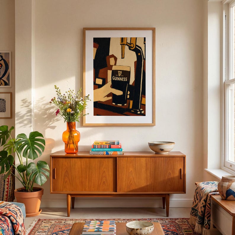 Op de afbeelding: Een ingelijste kunstafdruk met een Guinness-pint die wordt geschonken. Het kunstwerk heeft een vintage stijl met een hand die het glas vasthoudt. De afdruk hangt boven een houten kast met een oranje vaas en een plant.
