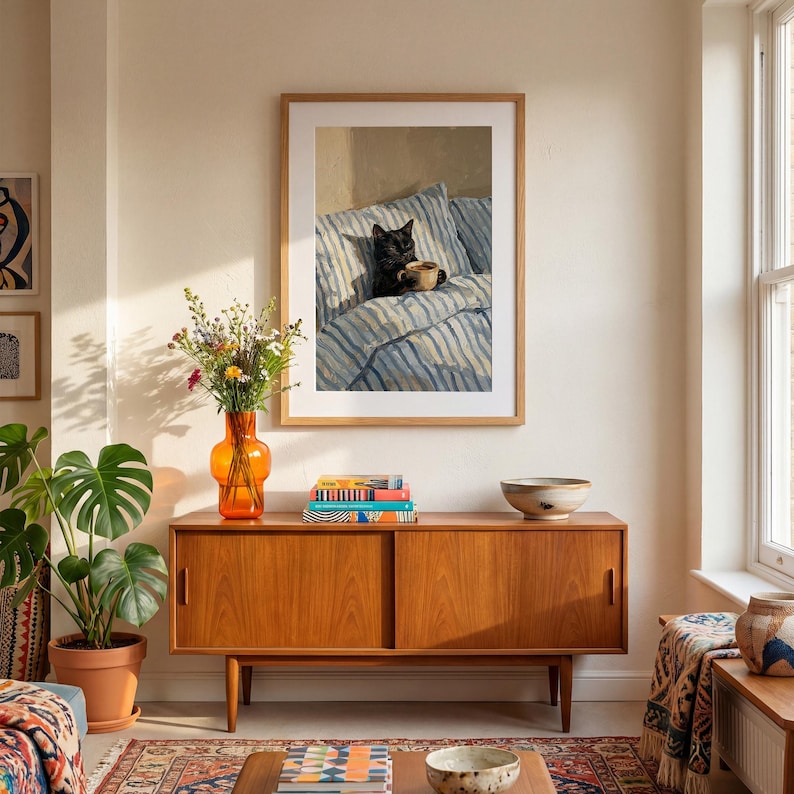 May include: A framed art print depicts a black cat holding a mug while lounging on a bed with blue and white striped bedding. The artwork is displayed above a wooden cabinet with an orange vase of flowers and a potted plant.