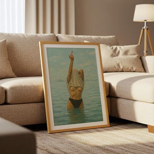 Puede incluir: Una impresi&oacute;n art&iacute;stica enmarcada que representa a una persona en el agua, levantando una camiseta. La obra de arte tiene una paleta de colores azul claro y beige, con un marco de madera. La imagen est&aacute; apoyada contra un sof&aacute; beige.