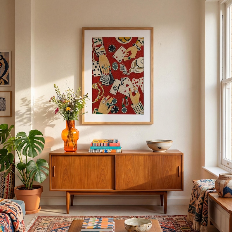 K&ouml;nnte beinhalten: Ein gerahmter Kunstdruck mit rotem Hintergrund, der H&auml;nde beim Kartenspielen, Pokermarker und Schmuck zeigt. Er h&auml;ngt &uuml;ber einem Holzschrank mit einer orangefarbenen Vase und einer Topfpflanze.
