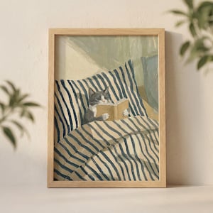 May include: A framed art print depicts a gray and white cat reading a book while lying on a bed with blue and white striped bedding. The artwork is in a wooden frame and has a soft, painterly style.