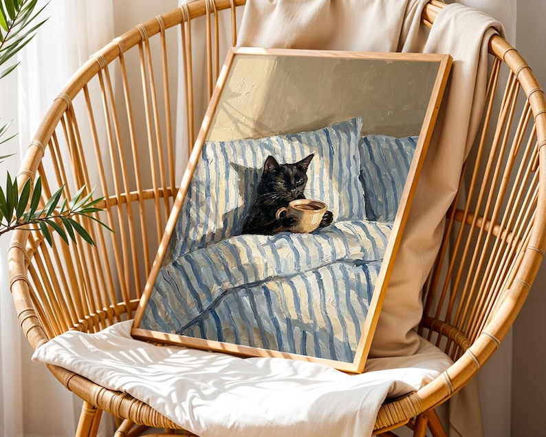 May include: A framed art print featuring a black cat holding a cup of coffee, resting on a bed with blue and white striped bedding. The artwork is in a wooden frame and is displayed on a wicker chair.