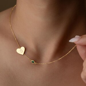 May include: A gold-colored necklace featuring a heart-shaped pendant with the letter "S" engraved on it. The necklace also includes a small green gemstone and is worn around the neck.