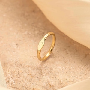 May include: A gold-colored ring with a smooth, polished finish. The ring features an oval-shaped face with the letter "T" engraved in a delicate script. The ring is set against a neutral background.
