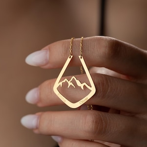 May include: A gold necklace featuring a mountain range within a diamond-shaped pendant. The pendant is inscribed with the names "Emma" and "Oliver". The necklace hangs from a fine chain.