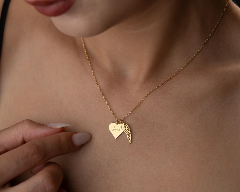 Puede incluir: Un collar dorado con un colgante en forma de coraz&oacute;n y un charm de ala. El colgante de coraz&oacute;n tiene la palabra "Love" grabada. El collar se lleva alrededor del cuello.