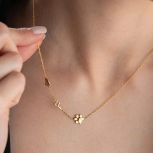 Op de afbeelding: Een goudkleurige ketting met een hartvormige bedel, de letters "LL" en een pootafdrukbedel. De ketting is zichtbaar op de huid van een persoon. De achtergrond is wazig, waardoor het ontwerp van de ketting en de details van de sieraden worden benadrukt.