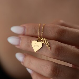 Puede incluir: Un collar dorado con un colgante en forma de coraz&oacute;n con el nombre "Lena" grabado y un dije de ala. El collar cuelga de una fina cadena. La joya est&aacute; sostenida por una mano.