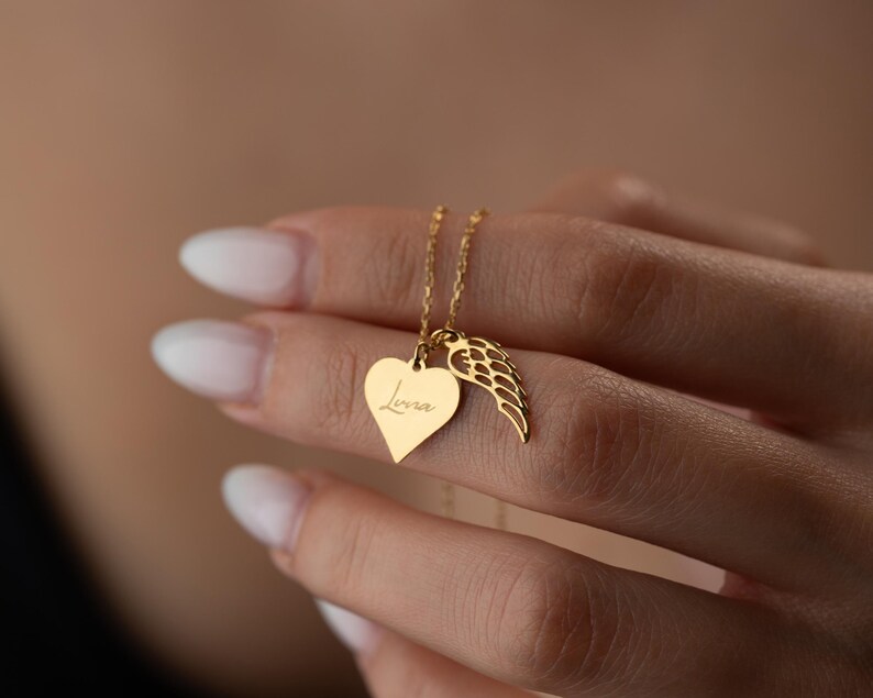 Puede incluir: Un collar dorado con un colgante en forma de coraz&oacute;n con el nombre "Lena" grabado y un dije de ala. El collar cuelga de una fina cadena. La joya est&aacute; sostenida por una mano.