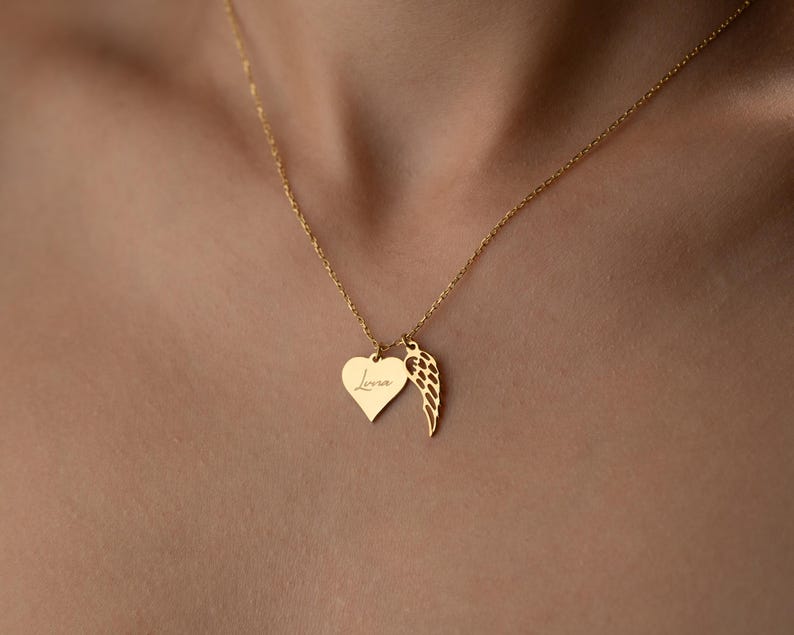 Puede incluir: Un collar dorado con un colgante en forma de coraz&oacute;n con la palabra "Love" grabada y un dije de ala. El collar se muestra en el pecho de una persona.