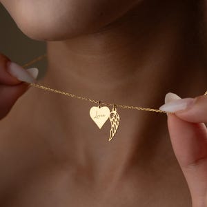 Puede incluir: Un collar dorado con dos colgantes: un coraz&oacute;n con la palabra "Love" y un ala. El collar est&aacute; sujeto por una persona. El colgante en forma de coraz&oacute;n mide aproximadamente 2 cm de ancho.