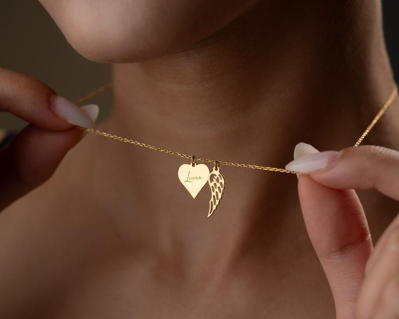 Puede incluir: Un collar dorado con dos colgantes: un coraz&oacute;n con la palabra "Love" y un ala. El collar est&aacute; sujeto por una persona. El colgante en forma de coraz&oacute;n mide aproximadamente 2 cm de ancho.