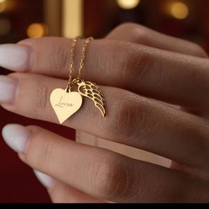 Puede incluir: Un collar dorado con un colgante en forma de coraz&oacute;n con el nombre "Luna" grabado y un charm de ala. El collar lo sostiene una persona. La cadena y los abalorios son de color dorado brillante. El colgante en forma de coraz&oacute;n mide aproximadamente 2 cm de alto.
