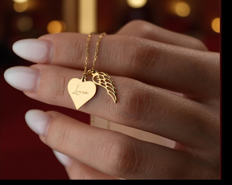 Puede incluir: Un collar dorado con un colgante en forma de coraz&oacute;n con el nombre "Luna" grabado y un charm de ala. El collar lo sostiene una persona. La cadena y los abalorios son de color dorado brillante. El colgante en forma de coraz&oacute;n mide aproximadamente 2 cm de alto.