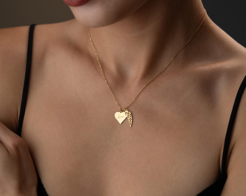 Puede incluir: Un delicado collar de oro con dos colgantes: un coraz&oacute;n y un ala. El colgante de coraz&oacute;n tiene una inscripci&oacute;n en cursiva. El collar se lleva sobre un fondo oscuro.
