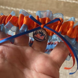 May include: A garter in orange and blue, featuring a Mets logo charm. The garter has a ruffled design with blue and orange ribbons. The charm is a silver-colored metal with the Mets logo.