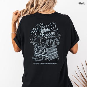 Pode incluir: Camiseta preta com um design gráfico branco nas costas. O design apresenta o texto "The Midnight Readers Book Club" com ilustrações de livros, velas e um relógio. O texto "Stories Over Sleep" e "Chasing Endings After Midnight" também estão incluídos.