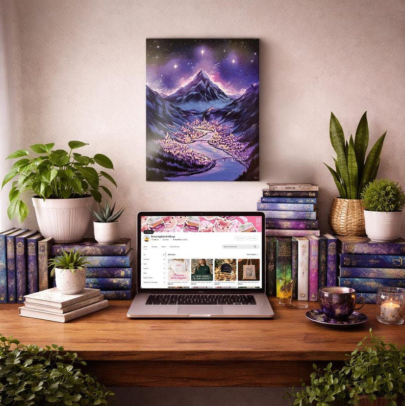 May include: A laptop displaying an Etsy shop, surrounded by plants, books, and a painting of a mountain landscape. The painting features a purple sky and a town nestled in a valley. A teacup and candle sit on the desk.